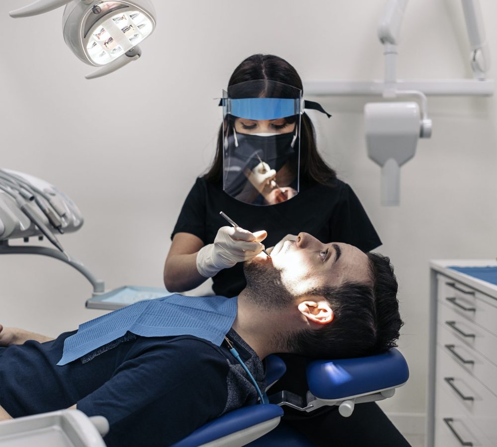 Female dentist wearing protective face mask working with young man during checkup revision.